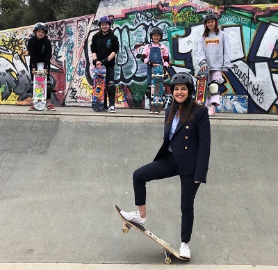 Hon Jane Howlett with a skateboard standing in front of a graphic wall. There are female skateboarders standing on top of the wall
