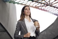 A young woman with long dark brown hair wearing a white blouse. She is holding her blazer and a coffee cup. She is standing under a building with a complex steel structure design