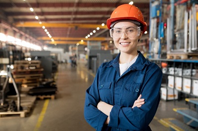 A young woman wearing an orange safety helmet. She is standing in a warehouse 